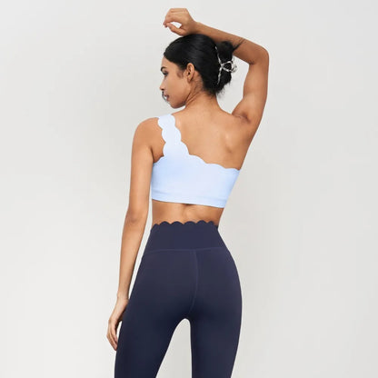 One-shoulder light blue yoga bra with scalloped edges, paired with high-waisted dark leggings, perfect for medium strength exercise.