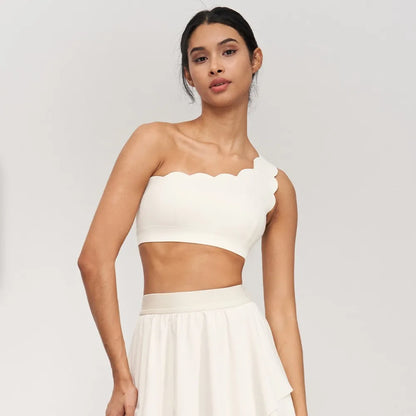 One-shoulder nude yoga bra with scalloped edges, designed for medium strength exercises, worn with a matching skirt.